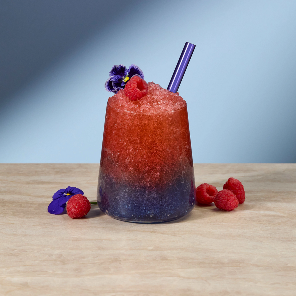 A tall, tapered glass filled with a red-to-purple gradient slushy drink, topped with raspberries, a purple edible flower, and a purple straw, set on a light wood surface against a blue-gray gradient background.