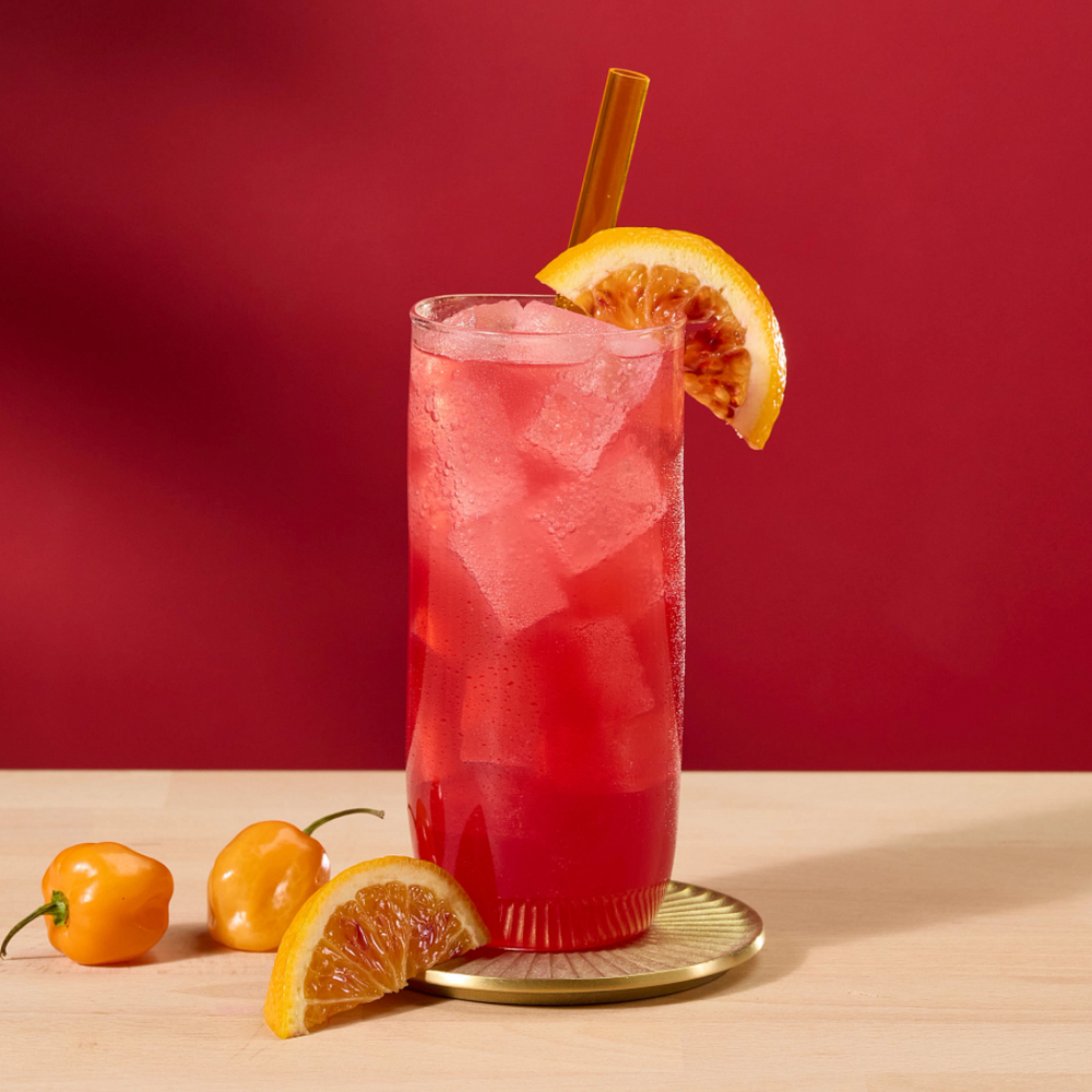A tall glass of red cocktail with ice, a blood orange slice garnish, and a straw, set on a light wood surface against a solid red background.