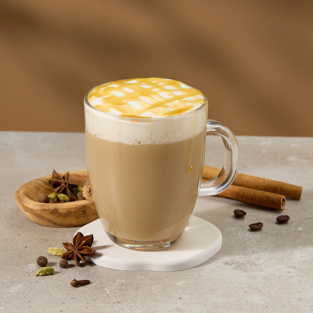 A glass mug filled with a latte or cappuccino topped with thick foam and a yellow grid pattern, set on a white coaster with cinnamon sticks, star anise, and coffee beans on a light stone surface against a warm brown background.