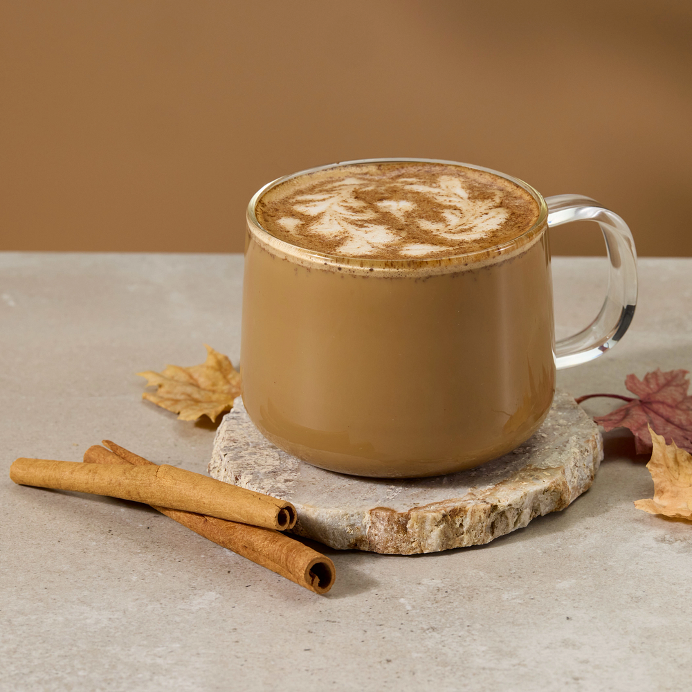 A clear glass mug filled with a layered coffee drink topped with latte art, set on a stone coaster with cinnamon sticks and autumn leaves on a beige surface against a warm brown background.
